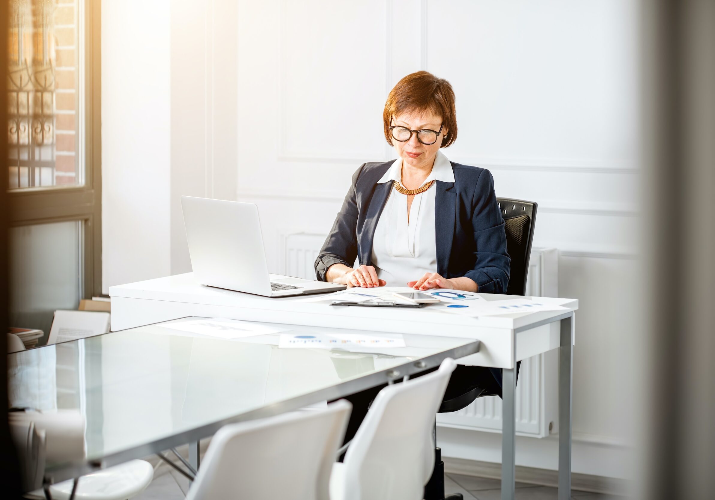 Elegant senior businesswoman dressed in the suit working with laptop and documents at the white office